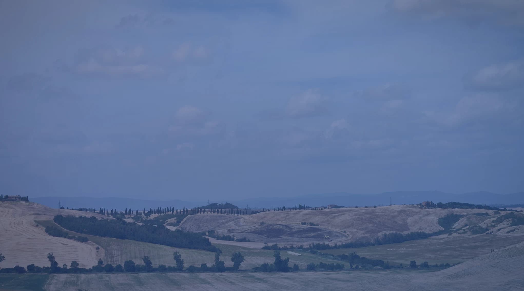 Rolling, partly harvested farm hills and fields under a wide, hazy summer sky, like the open country of the central plains.