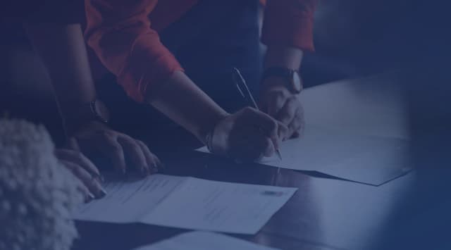 Two people at a table, one signing loan paperwork with a pen while the other holds the pages steady
